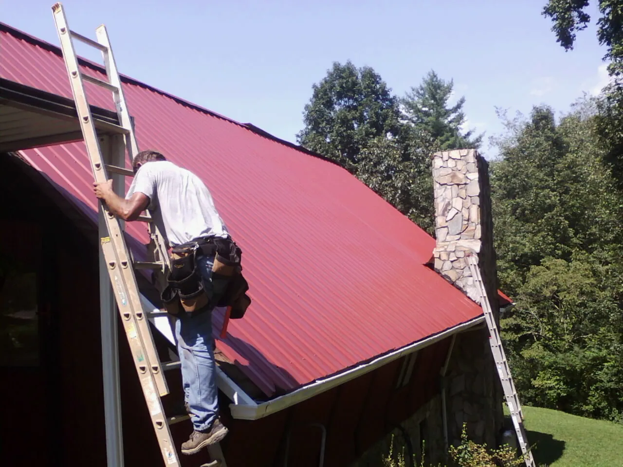 Roofer on a ladder beside a red metal roof and stone chimney — Soffit & Fascia Repair by Roofing Brothers in Brookston